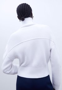 Vit stickad tröja med hög krage, kortad design och ribbad fåll. Har texturerade, vertikala linjer och sänkta axlar.