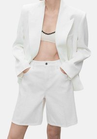 Blazer blanc surdimensionné avec épaulettes, bouton unique, associé à un short blanc taille haute. Texture lisse, design minimaliste.