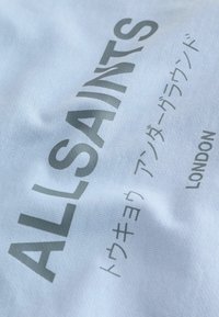 Tissu blanc avec texte gris indiquant "ALLSAINTS", caractères japonais en dessous, et "LONDON" aligné verticalement sur le côté droit.