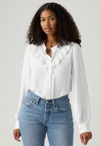 Camicia bianca con bottoni realizzata in un tessuto leggero, caratterizzata da un bordo in pizzo arricciato lungo il collo, abbinata a jeans in denim blu a vita alta.