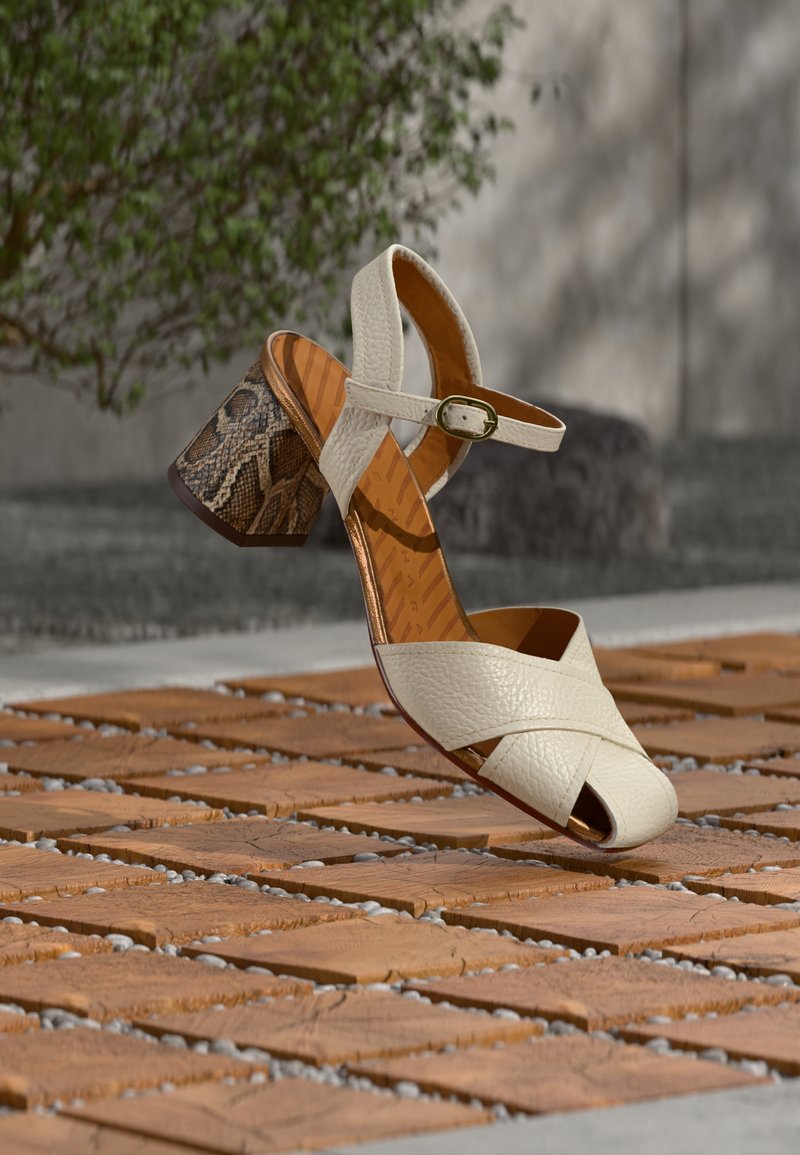 Άσπρο δερμάτινο σανδάλι με λουριά που διασταυρώνονται και αγκράφα, με χοντρό τακούνι σε σχέδιο δέρματος φιδιού, τοποθετημένο πάνω σε πλακόστρωτο από τούβλα.