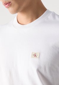 Primo piano di una persona che indossa una maglietta bianca a girocollo con una piccola toppa beige raffigurante le lettere rosse "CK" sul petto.