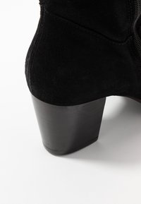 Bottine en suede noire avec un talon épais noir. Présente une texture lisse et une couture visible le long du côté.