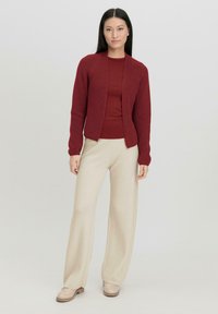 Roter gestrickter Cardigan über einem passenden Oberteil, kombiniert mit beigen Schlaghose und hellen Schuhen, vor einem neutralen Hintergrund.