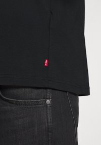 Czarny dół koszuli Levi's z czerwoną metką logo na tle ciemnoszarych dżinsów, widok z boku z bliska.