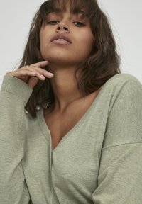 Maglione verde chiaro a V con una texture morbida, spalle basse e maniche lunghe. Il tessuto appare liscio e leggero.