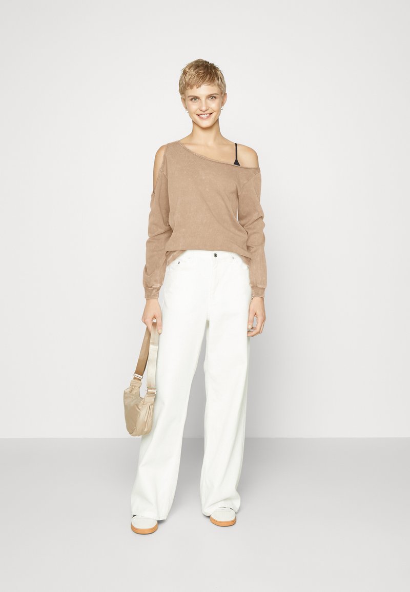 Hellbraunes Off-Shoulder-Oberteil, weiße weit geschnittene Hose und beigefarbene Handtasche. Das Modell trägt lässige weiße Sneakers; minimalistischer Stil mit lockerer Passform.