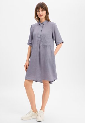 Femme portant une robe chemise grise ample jusqu'aux genoux, manches retroussées et baskets blanches, debout avec une main dans la poche sur fond uni.