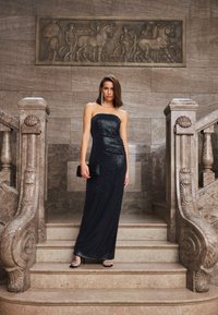 Abito nero senza spalline con silhouette aderente e texture di paillettes. La modella tiene una pochette nera e indossa sandali con tacco aperti e cinturini su una scalinata di marmo.