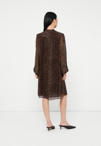 Sieviete ar gariem melniem matiem, tērpusies ceļa garumā, garām piedurknēm caurspīdīgā brūnā leopardu apdruku kleitā un melnās kaķu papēžu mules, stāv ar muguru pret skatītāju.