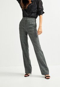 Pantalon à carreaux gris avec une taille haute et des jambes droites, fabriqué dans un tissu texturé. Associé à une chemise noire boutonnée et des talons ouverts.