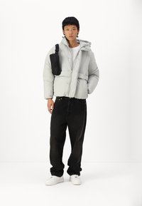 Chaqueta de plumas gris claro con capucha, con cremallera frontal y botones a presión; combinada con jeans negros holgados y un pequeño bolso negro de hombro.