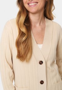 Šviesiai rudas juostuotas kardiganas su ilgomis rankovėmis, V formos iškirpte ir dviem priekinėmis kišenėmis. Funkciniai tamsiai rudi mygtukai ir minkštas audinys.