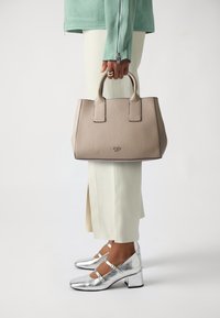 Beige textured leather handbag with two short handles, rectangular shape, and embossed logo. Worn with light-colored trousers and silver shoes.