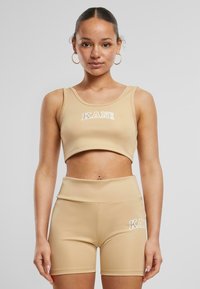 Beige sportsettet inkluderer en kort tanktopp og høye shorts, begge med en glatt tekstur og "KANI"-logo i hvitt.