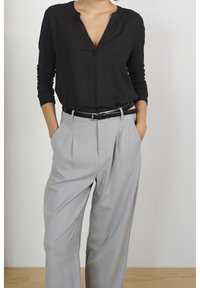 Kvinde iført en sort langærmet skjorte, stoppet ind i lysegrå plisserede bukser med sort bælte, hænderne i lommerne, stående op ad en hvid væg.