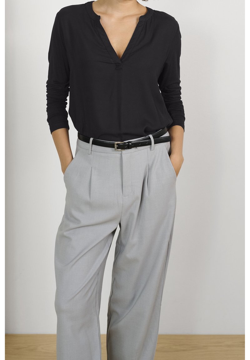 Kvinde iført en sort langærmet skjorte, stoppet ind i lysegrå plisserede bukser med sort bælte, hænderne i lommerne, stående op ad en hvid væg.