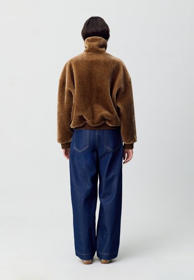 Sudadera de forro polar marrón, suave y de cuello alto, combinada con pantalones vaqueros de mezclilla oscuros y holgados, que presentan bolsillos traseros y detalles de costura visibles.