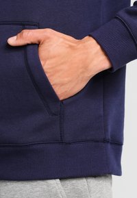 Felpa blu navy con polsini e orlo a costine, dotata di tasca a marsupio. Il tessuto appare morbido con una texture liscia.