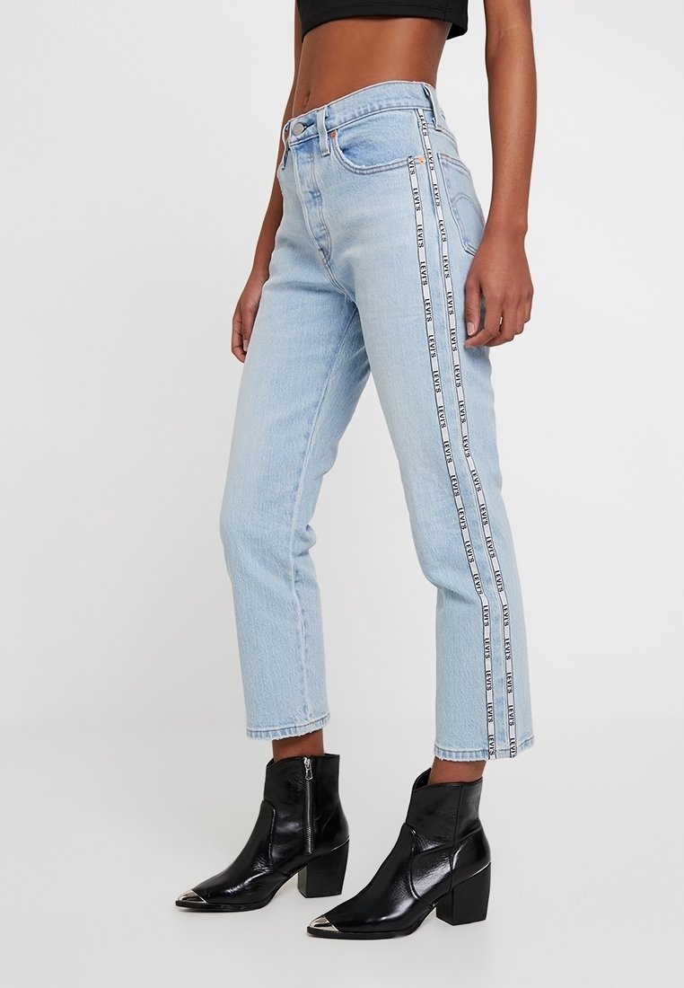 Jean taille haute bleu clair avec une coupe droite, présentant des bandes latérales avec du texte. Associés à des bottines noires. Texture en denim lisse.