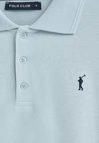 Ljusblå polo med texturerad tyg, tre knappar och en liten broderad golfspelare i marinblått på vänster bröst.