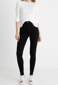 Top blanco de manga larga con diseño texturizado, combinado con jeans ajustados negros de tiro alto y zapatillas blancas con acentos plateados.