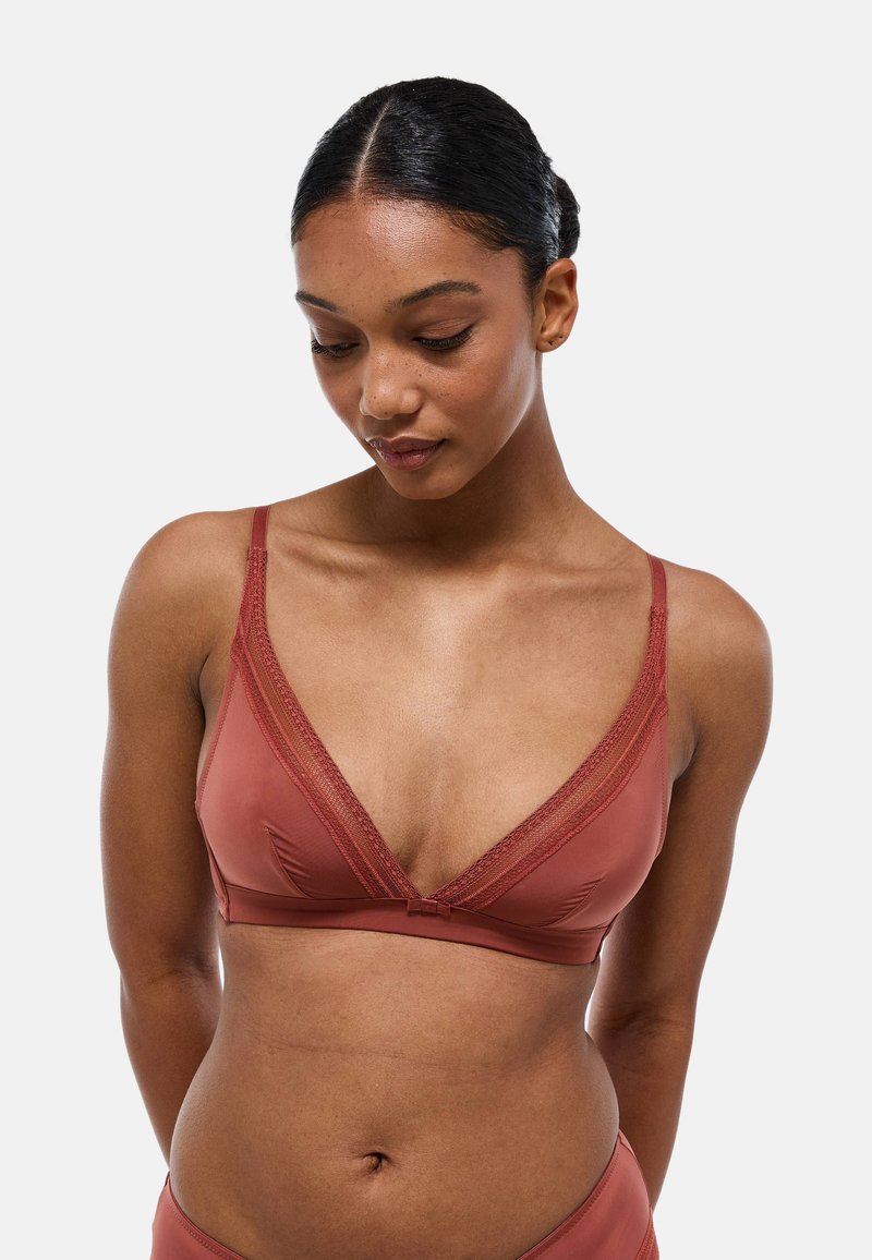 Soutien-gorge de couleur rouille en tissu doux et lisse. Il présente un décolleté plongeant, des finitions en dentelle délicate sur les bords, et des bretelles ajustables.