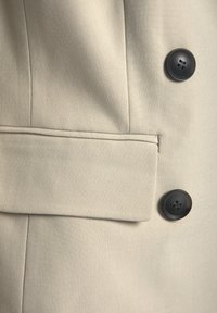 Manteau en tissu beige avec deux boutons foncés et une poche à rabat horizontale sur le panneau avant.