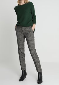 Maglione verde strutturato con scollatura a barca, abbinato a pantaloni a quadri bianchi e neri e stivaletti neri alla caviglia.