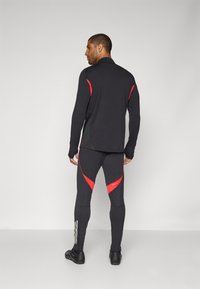 Zwarte sporttop en tights met rode accenten. Lange mouwen, ronde hals en een getailleerd ontwerp. Textuurstof met reflecterende strepen op de tights.