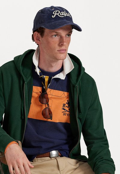 Jovem vestido com um boné azul "Ralph", um hoodie verde com fecho éclair, uma camisa de rugby às riscas com óculos de sol pendurados, calças caqui e um cinto castanho, sentado contra um fundo branco.