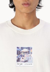 Hvid sweatshirt med et trykt foto af en gadesælger-vogn med en paraply og skilt, overlay-tekst lyder "New York City of Dreams."