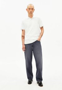 Wit poloshirt met korte mouwen, gecombineerd met donkerblauwe, loszittende spijkerbroeken en zwarte schoenen. Tatoeages zichtbaar op de rechterarm.