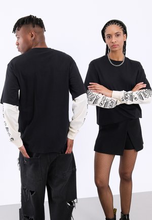 Czarny, oversize T-shirt z długimi kremowymi rękawami z czarnym nadrukiem. Model nosi czarne, podarte spodnie oraz rajstopy z siateczki. Prosty design.