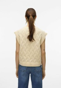 Beige gebreide mouwloze trui met een textuur kabelpatroon, geribde zoom en relaxte pasvorm, gecombineerd met blauwe high-waisted jeans.