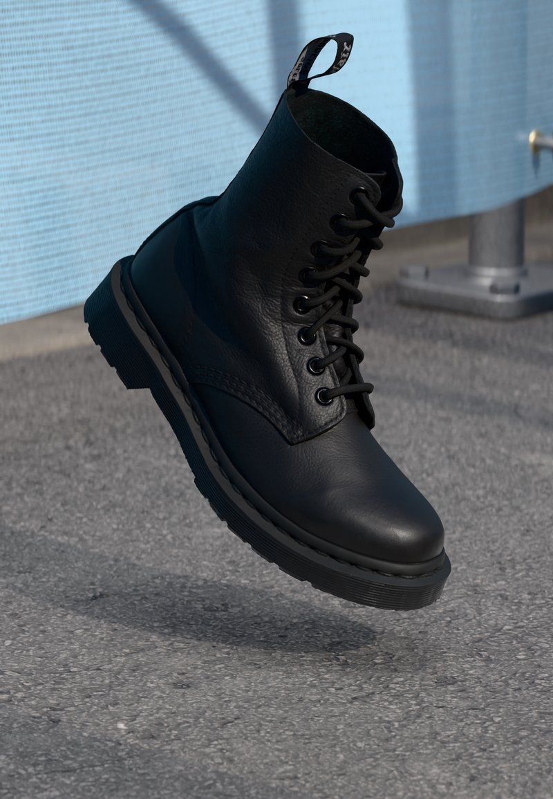 Botte noire en cuir à lacets suspendue en l'air au-dessus d'un sol pavé gris avec un mur bleu et un poteau métallique en arrière-plan.