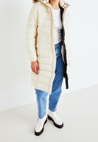 Beżowy płaszcz ocieplany z kapturem, z zamkiem błyskawicznym, bocznymi kieszeniami i poziomym pikowaniem. Nosi się go na białą bluzkę i niebieskie dżinsy.