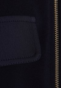 Detail ze tmavě modré látky s hladkým povrchem, obdélníkovou kapsou se šitými okraji a zlatým zipem na boku.