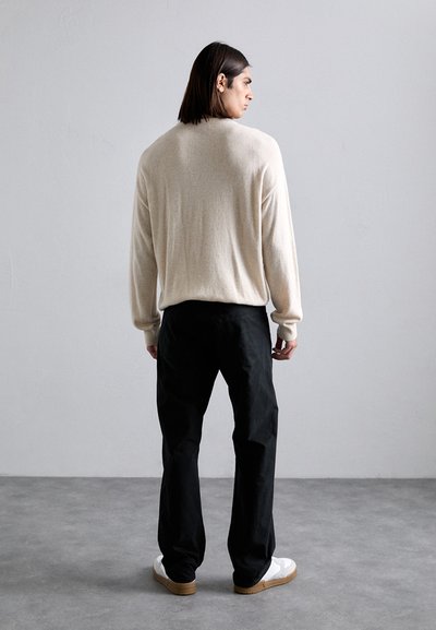 Suéter beige de punto con mangas largas, combinado con pantalones negros y zapatillas blancas. La textura parece suave y el corte es relajado.