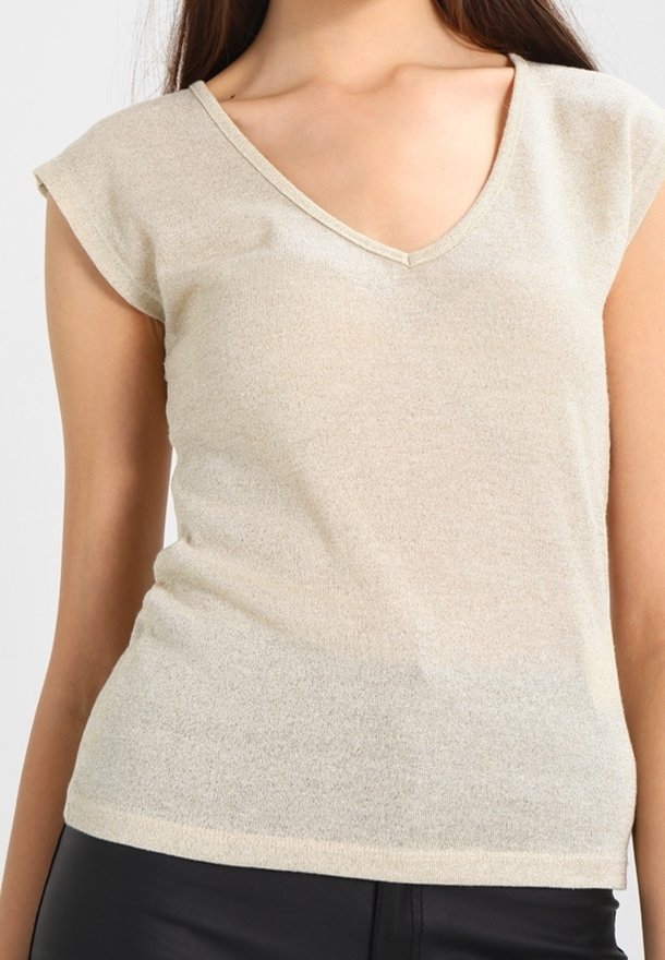 Haut beige à manches courtes avec col en V porté par une personne aux longs cheveux foncés, associé à un pantalon noir sur un fond clair uni.