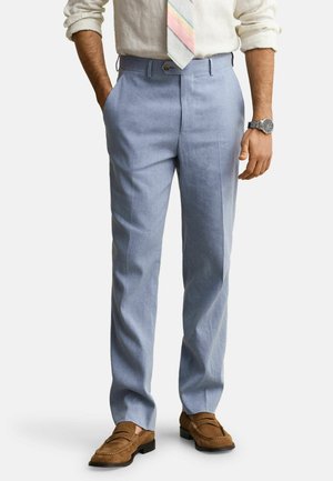 Un homme porte un pantalon habillé bleu clair, des mocassins marron clair, une chemise blanche à manches longues retroussées, une cravate à rayures pastel et une montre-bracelet argentée, les mains détendues.