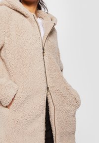 Manteau beige à capuche à texture duveteuse, fermeture à double zip, porté par-dessus un haut blanc, mains dans les poches.