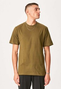 Ung mand med kort hår iført olivengrøn t-shirt, sorte bukser, sølvhalssmykke og armbånd, der kigger til siden mod en ensfarvet baggrund.