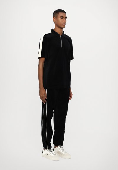 Polo noir en velours à manches courtes avec une fermeture éclair quart de zip et des rayures blanches sur les épaules, associé à un pantalon noir assorti et des baskets blanches.