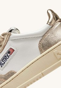 Sneaker bianca con accenti metallici dorati, lati texturizzati e un tab sul tallone. Presenta una patch con logo e una suola in gomma beige con un battistrada minimale.