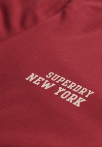 Rødt bomuldsstof med en blød tekstur, der har hvid trykt tekst "SUPERDRY NEW YORK" i en blokagtig skrifttype i midten.