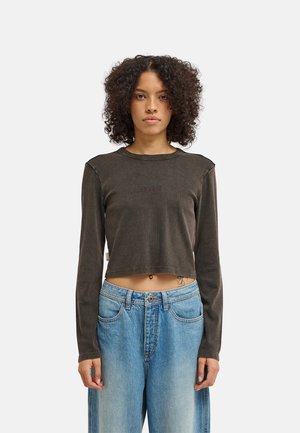 Persona con capelli ricci che indossa un crop top scuro a maniche lunghe e jeans blu larghi sta in piedi rivolta in avanti su uno sfondo chiaro e uniforme.