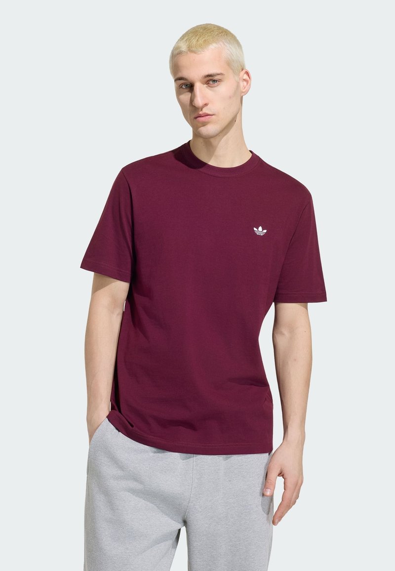 Jeune homme aux cheveux blonds courts portant un t-shirt uni bordeaux avec un petit logo blanc et un pantalon de survêtement gris clair, debout de manière détendue.