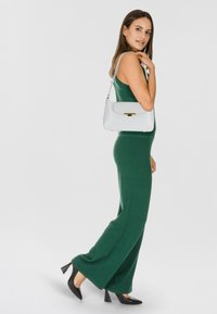 Jumpsuit verde senza maniche abbinato a una borsa bianca con chiusura dorata. Il modello indossa scarpe con tacco nere. Sfondo minimale.