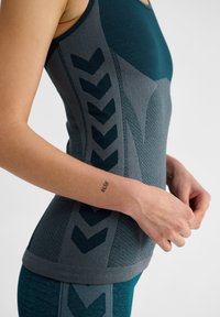 Teal sporttop met een gestructureerd grijs zijpaneel en donkerblauwe chevronpatronen, gemaakt van een rekbare, ademende stof.